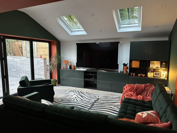 Modern living room with green walls, large windows, a dark TV unit, and a colourful sofa.